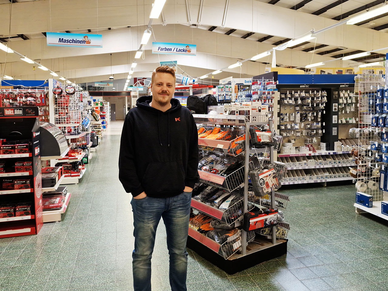 Ein Mitarbeiter in schwarzem Hoodie steht in einem Baumarkt vor Regalen mit Werkzeugen und Zubehör. Ein Mitarbeiter in schwarzem Hoodie steht in einem Baumarkt vor Regalen mit Werkzeugen und Zubehör.